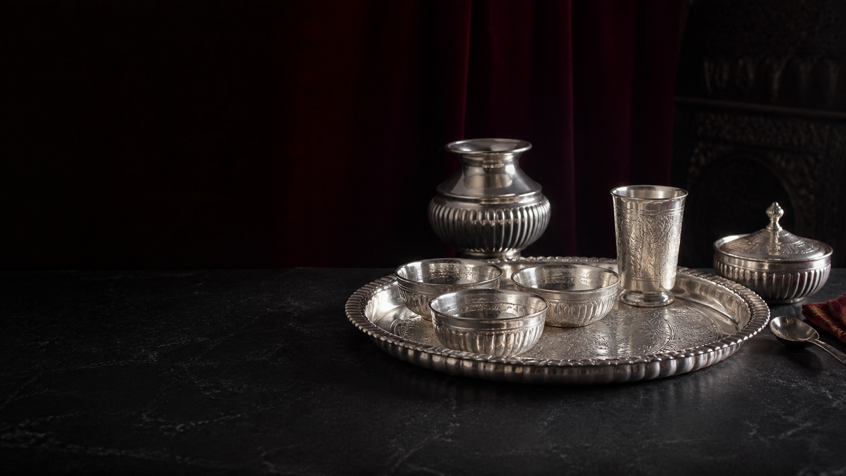 Silver thali and serving pieces arranged on a dark stone table